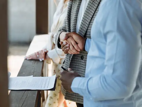Two people shaking hands over a wooden ledge next to a signed paper and a pen, in warm outdoor daylight