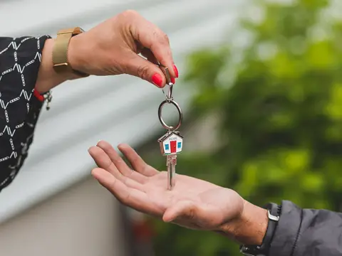A hand lowering a single house key on a small house-shaped keychain into another person's open palm, with greenery blurred behind