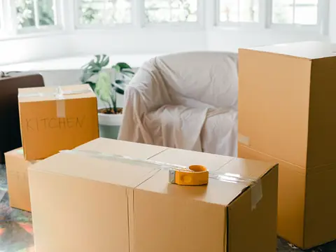 Stacked cardboard moving boxes, one labeled KITCHEN by hand, with a tape dispenser, a suitcase and a covered armchair in a bright room