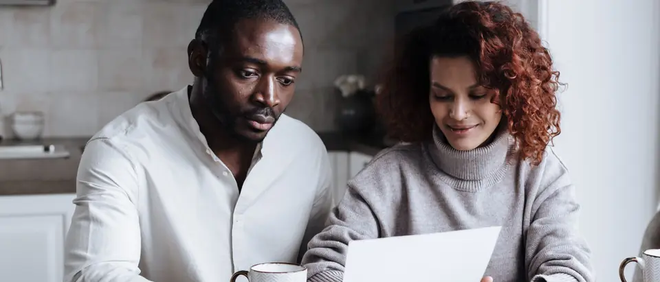 A couple sitting at a kitchen table calmly reviewing a sheet of paper, with coffee mugs, a pen, documents and a laptop in front of them